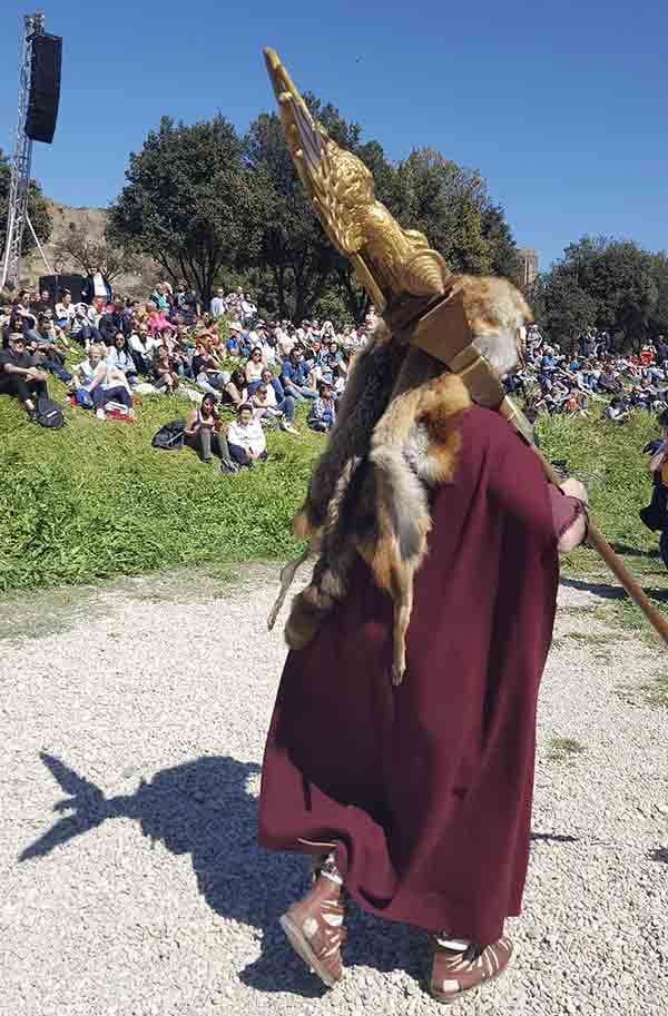 An actor dressed in replica ancient Roman clothing carries an eagle standard in front of a crowd.